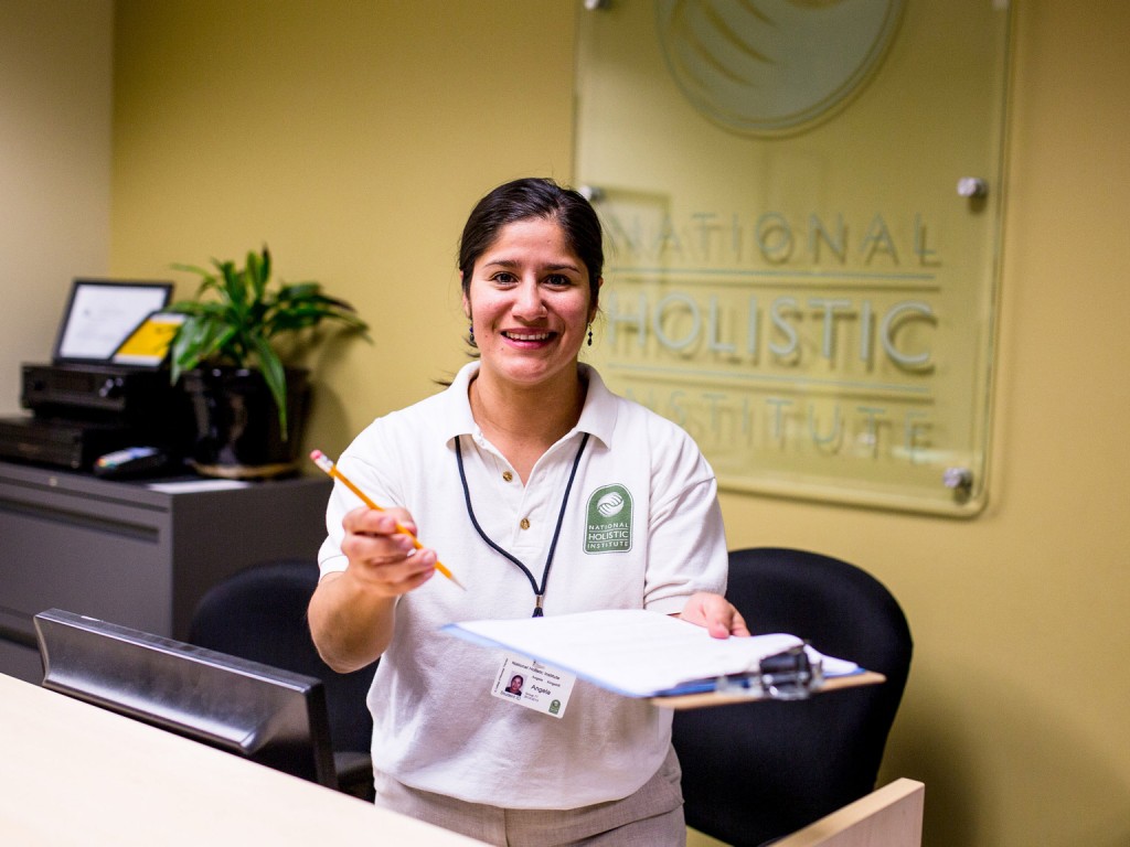 An NHI massage school staff member standing behind a desk at the Admissions office and holding a pencil and a paper offering you to sign