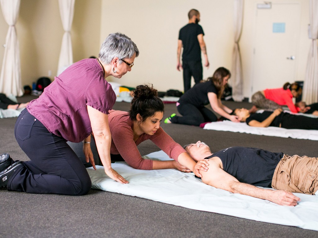 A woman giving a Shiatsu massage to a person lying in a pad on the floor while a professor is supervising