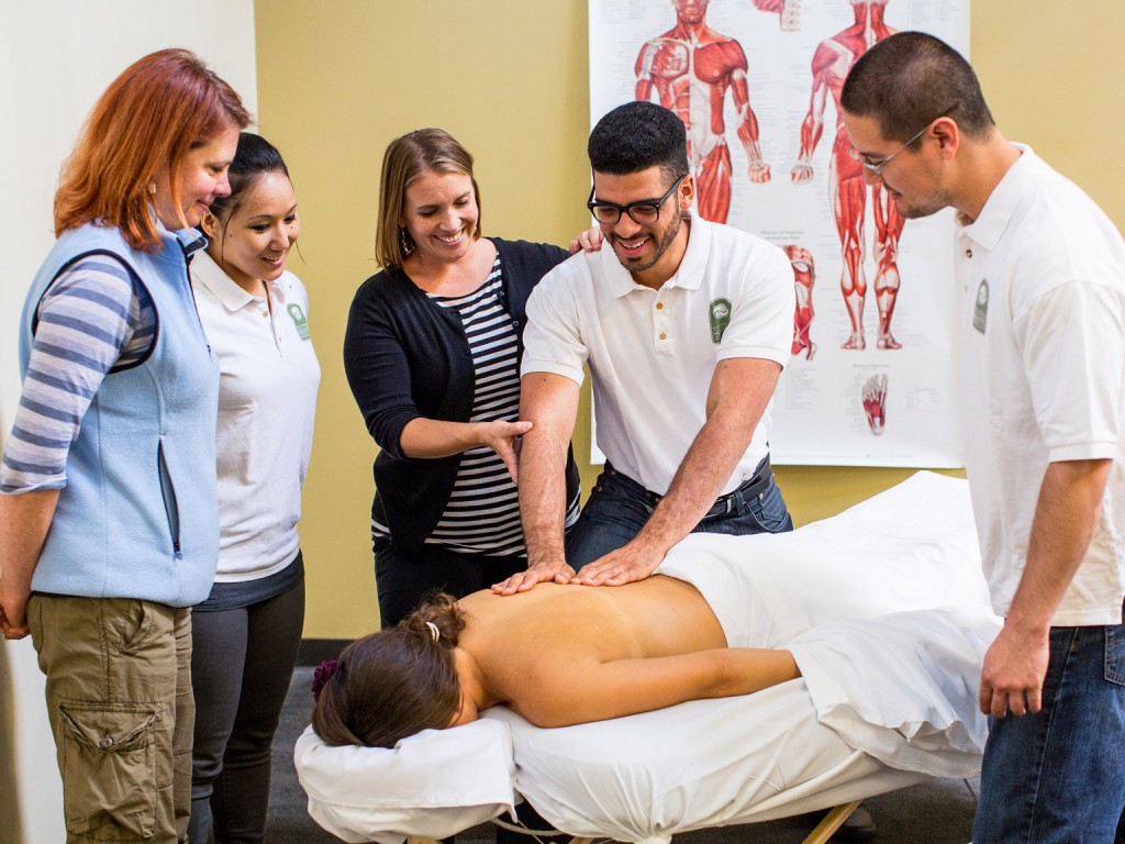 An NHI massage school student giving a back massage to a person laying at a massage table while two students and two professors watch and smile.